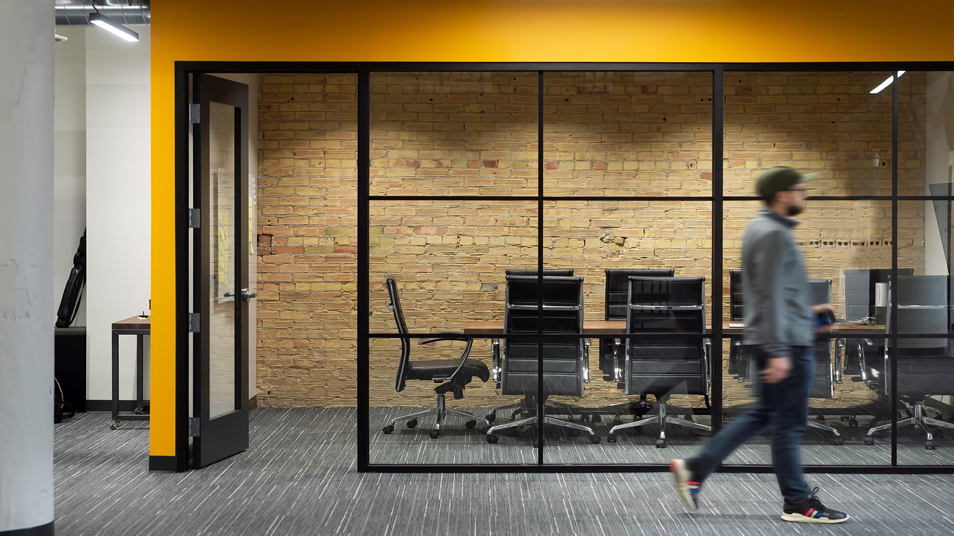 A man walks past a meeting room with a yellow wall in the Aptera Office.