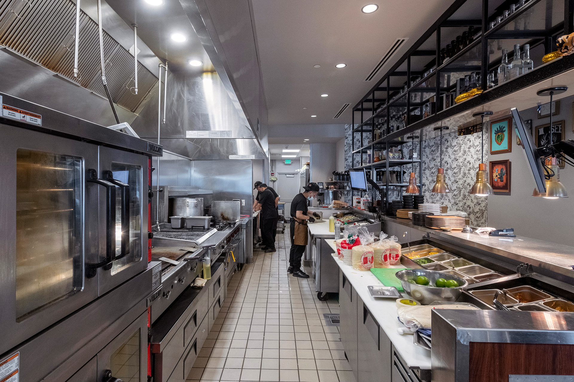 A section of the kitchen is visible to guests, allowing them to be part of the creative cooking and dining experience. Sleek equipment, decorative shelving, and deliberate lighting design blend the kitchen and dining spaces.