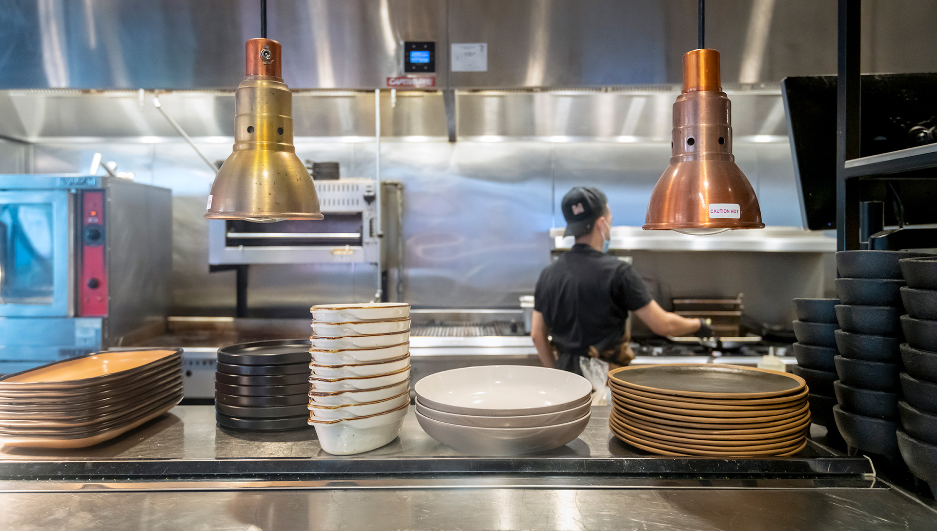 A section of the kitchen is visible to guests, allowing them to be part of the creative cooking and dining experience. Sleek equipment, decorative shelving, and deliberate lighting design blend the kitchen and dining spaces.