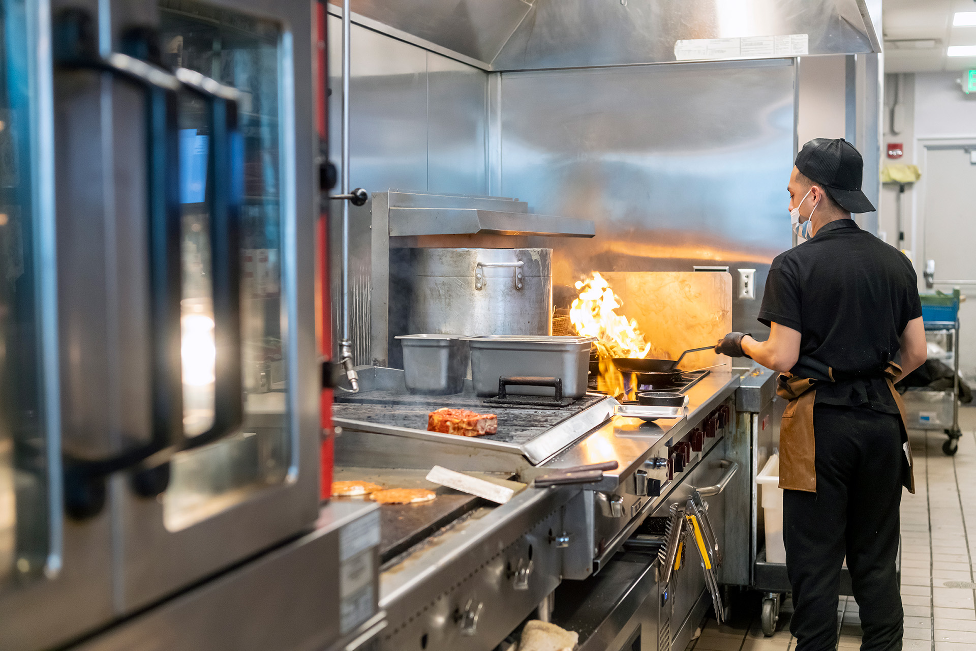 A section of the kitchen is visible to guests, allowing them to be part of the creative cooking and dining experience. Sleek equipment, decorative shelving, and deliberate lighting design blend the kitchen and dining spaces.