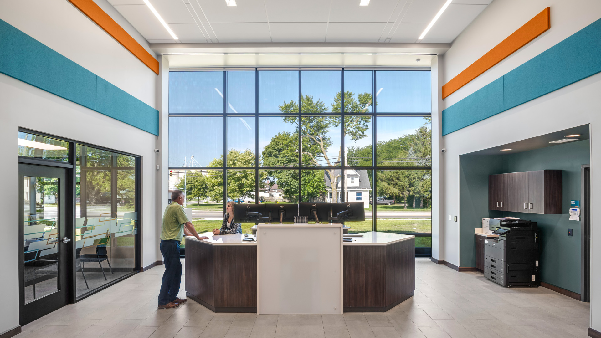 Teller line in bank branch with bright orange and blue branding and large windows.