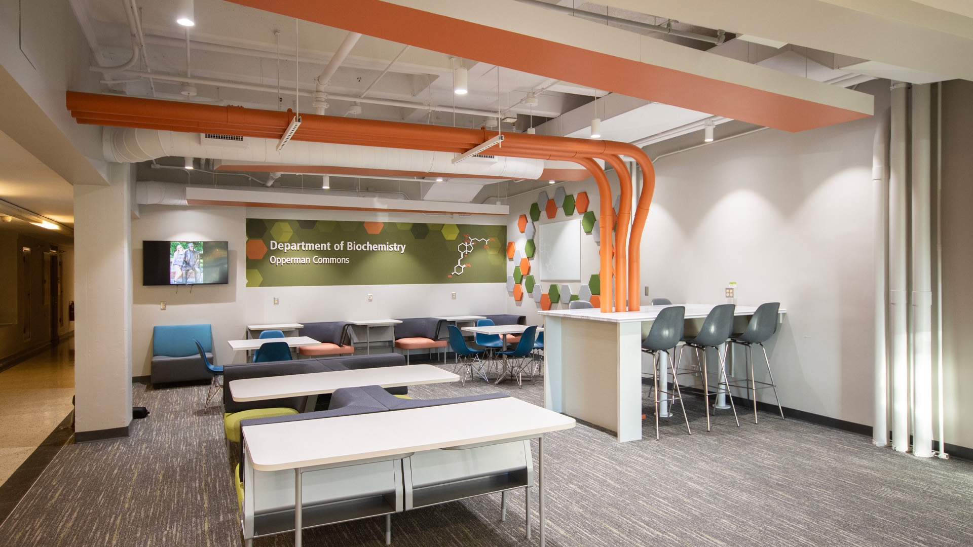 Student hub with bold orange, green, blue, and gray seating, pipes, and tables.